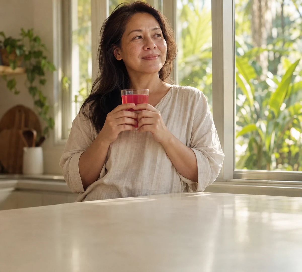Filipina woman in her 40s holding a glass of pink drink by a sunlit kitchen window in the morning